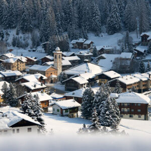 Snow-covered chalets and a church with a bell tower nestle in a wooded alpine valley under low cloud.