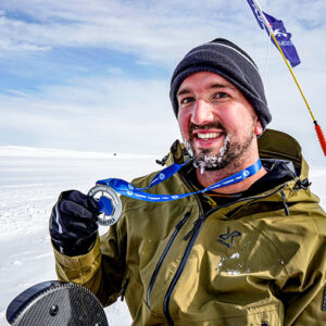 A smiling, frost-bearded man in a green jacket and black beanie holds up a medal in a vast snowy landscape beneath a partly cloudy sky.