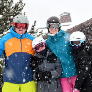 Four skiers in helmets and goggles pose together in the snow, with two adults in bright jackets and two children in darker coats.