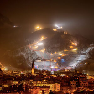Kitzbühel Unveils a Winter Wonderland of Sporting Spectacles and Culinary Delights Floodlit ski slopes descend through mist above a warmly lit alpine town, its church spire rising among snow-dusted rooftops.