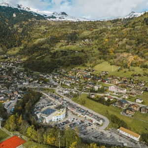 Verbier Tourisme introduces multi mode of transport booking service Aerial view of an alpine village with chalets, a supermarket and parked cars, set against forested slopes and snow-capped peaks. (124 characters)