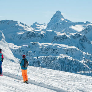 Vialattea partners with Trenitalia to encourage sustainable ski travel Two skiers with backpacks pause on a snowy slope, gazing towards a dramatic pointed peak under clear blue skies.