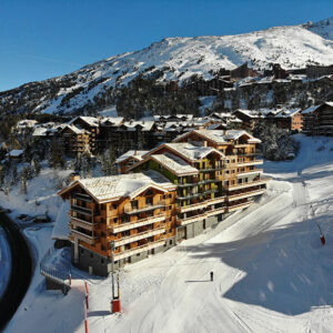 Snow-covered wooden chalet buildings cluster on a mountainside, with a lone skier on the piste below and snow-capped peaks rising under a clear blue sky.