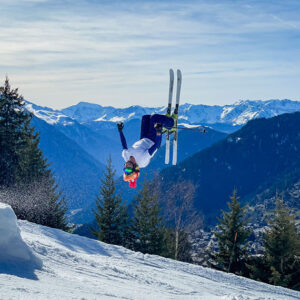 Boss des Bosses returns to Verbier, Switzerland A skier performs an upside-down aerial trick above a snowy slope, with snow-capped mountains and a valley town below.