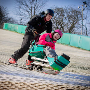 Travel agency's fundraising for DSUK increases almost tenfold An adult in black ski gear assists a child wearing a pink helmet, seated in a green adaptive ski sled, on a gentle snow slope lined with green barriers.