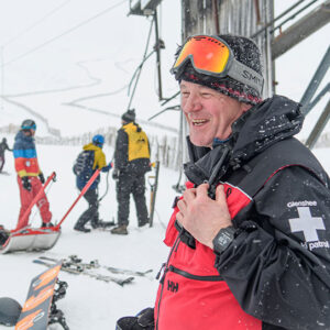 Celebrating ski patrollers worldwide on International Ski Patrol Day 2024 Smiling ski patroller in a red and black Glenshee jacket with goggles on his beanie, standing on a snowy slope with other skiers and a ski lift behind him.