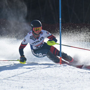 A slalom skier in a dark racing suit and VITALINI bib carves aggressively around a blue gate, sending snow spraying into the air.