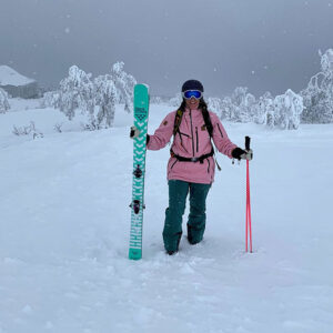 Person in a pink jacket and goggles stands in deep snow holding a teal ski upright, with snow-laden trees and a building behind.