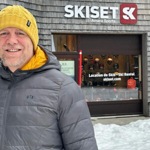 Mike Tindall's latest charity challenge takes to the slopes A smiling man in a yellow beanie and grey puffer jacket stands outside a Skiset ski rental shop with snow around.