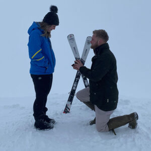 Skier pops the question on mountain top despite injury A man kneels in the snow presenting a small box to a woman in a blue jacket, skis propped up between them.