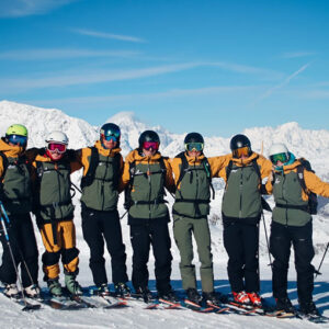 New junior ski camps for kids in Tignes Seven skiers in matching yellow jackets, helmets and goggles stand arm-in-arm on a snowy slope with mountain peaks behind them.