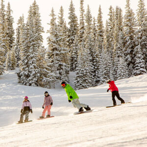 Last-minute deal for a family-friendly adventure in Aspen Snowmass Four snowboarders in colourful jackets descend a snowy slope lined with snow-laden evergreen trees.
