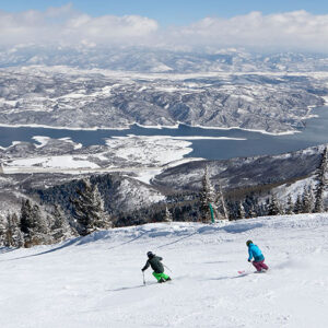 Get inspired ahead of Ikon Pass going on sale for 24/25 season Two skiers carve down a snowy mountainside above a frozen lake and snow-patched peaks under a partly cloudy sky.