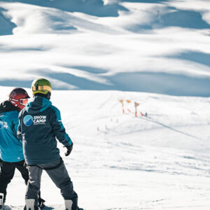 Snow sports inclusion initiative helping to turn young lives around Two snowboarders in helmets and winter gear stand on a sunlit slope, with ski lifts visible in the distance.