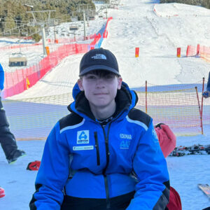 A young man in a blue Colmar ski jacket and black The North Face cap sits in the snow using a laptop, with a ski slope and red safety netting behind him.