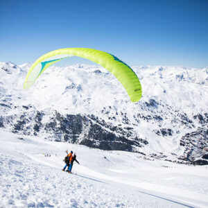 A bird’s eye view of Meribel A skier with a bright yellow paraglider soars above a snowy alpine slope with distant mountain ranges under a clear blue sky.