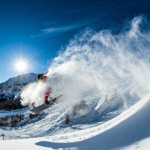 A snowboarder launches off a powder-covered slope, sending up a huge spray of snow against a bright blue sky and shining sun.