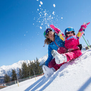 A woman and child in bright ski gear laugh as they throw snow into the air on a sunny mountain slope.