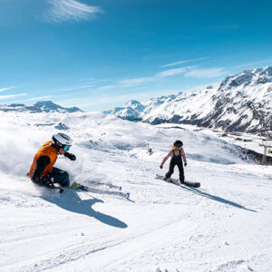 Ikon Pass adds St. Moritz for Winter 2024/25 A skier in an orange jacket carves through fresh powder ahead of a snowboarder on a sunny mountain slope with distant peaks and a chairlift visible.