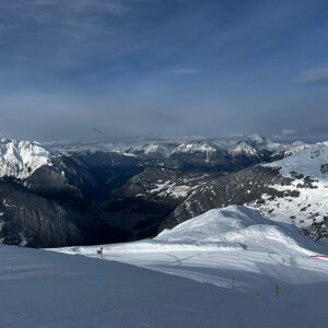 Verbier's Alpine Ascent Towards Eco-Friendly Excellence A vast snow-covered mountain range with a deep forested valley, distant jagged peaks under a partly cloudy sky, and two tiny skiers on the groomed slope.