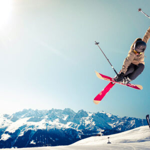 Skiers 'willing to pay 11% more for an eco-friendly snowsports holiday' A skier in a tan jacket and helmet launches into a bright blue sky, skis crossed mid-jump above snow-capped mountain peaks.