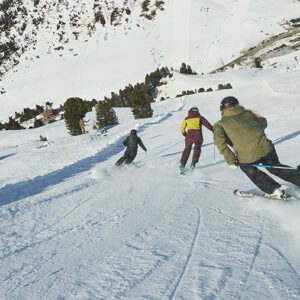 Ski Club of Great Britain reveals latest trends Three skiers carve down a snowy mountain slope, kicking up powder with a ski resort and treeline visible in the background.