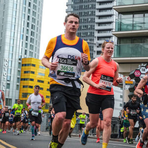 Runners in numbered race bibs jog down a city street lined with tall glass buildings, led by a man in a blue and yellow vest and a woman in a coral tank top.