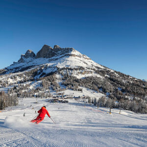 Champion's race to success at Italian boutique-style ski resort A skier in red carves down a snowy slope, kicking up powder, with a rocky mountain peak and forested valley behind under a clear blue sky.