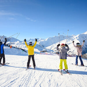 Four skiers in colourful jackets stand on a snowy slope, raising their poles triumphantly towards snow-capped peaks under a clear blue sky.