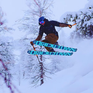 Carve Your Way To Panorama With Frontier Ski & Air Canada A skier in dark gear and blue goggles launches through deep powder on turquoise Black Crows skis between snow-laden trees.