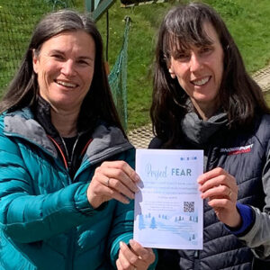 Snowsports Fear Surveyed To Reduce Barrier To Snowsport Enjoyment Two smiling women stand together holding a Project FEAR leaflet, one in a teal puffer jacket and the other in a dark Snowsport branded gilet.