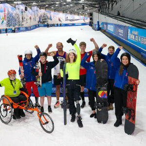 A group of ten people, including a person in a wheelchair, pose cheerfully on an indoor ski slope, holding snowboards and ski poles near blue Snozone barriers.