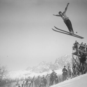 In the footsteps of the first Winter Olympians A ski jumper soars high through the air with one arm raised above spectators and a wooden viewing tower, against snow-capped mountains.