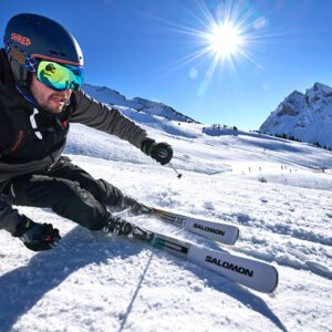 A skier in a black Shred helmet and green goggles carves down a sunlit slope on white Salomon skis with snowy mountains behind.