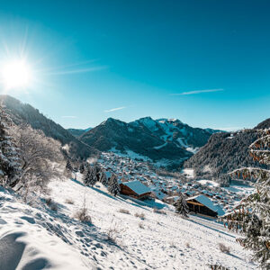 Snow-covered wooden chalets sit in a sunlit valley beneath towering peaks, framed by frost-laden fir trees.