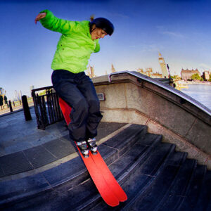 Epic ski rail jam set to launch UK flagship store Skier in a green jacket and helmet grinds a handrail on red skis down stone riverside steps with a city skyline behind.