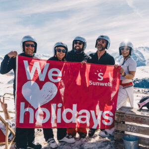 Tomorrowland Winter returning for 2025 Five people in ski helmets and sunglasses hold a red "We love Sunweb holidays" banner on a snowy mountain.