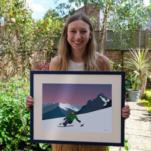 Artist's series of adaptive snowsports designs raising funds for DSUK A smiling woman with long hair stands in a leafy garden holding a framed illustration of a skier on a snowy mountain slope at dusk.