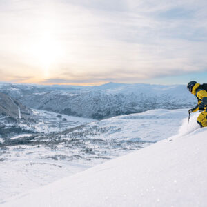 Free Private Lessons in Norway on offer with Ski Safari A skier in a yellow jacket carves down a steep snow-covered mountain slope above a vast wintry valley at sunset.