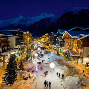 A snow-covered alpine village glows with warm light from wooden chalets and spherical street lamps as pedestrians wander beneath snow-capped mountains at dusk.