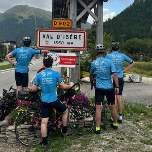 Team celebrates completing charity bike ride to Val d'Isère Four cyclists in blue jerseys and helmets pose with their bikes beside a Val d'Isère 1850m road sign, with mountains in the background.