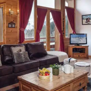 Catered vs self-catered chalets – which is best for you? Cosy wooden chalet living room with brown leather sofas, red curtains, a wood-burning stove, and snowy mountain views through the windows.