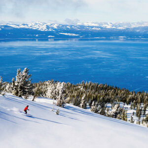Savings to be made on trips to North America says Tour Operator A snowboarder in a red jacket carves down a snowy slope above a vast blue lake backed by distant snow-capped mountains.