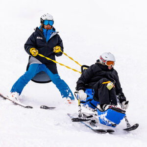 Award win for Disability Snowsport UK A standing skier uses yellow outriggers to guide a seated sit-skier down a snowy slope.