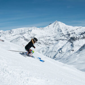 Val d'Isère’s Slopes Set To Open After Fresh Snowfall Two skiers carve down a sunlit slope with jagged snow-capped peaks rising behind them under a clear blue sky.