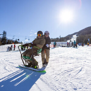 Two helmeted skiers pose on a sunny slope, one seated in an adaptive sit-ski, with chairlifts and other skiers visible behind them.