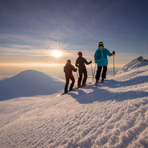 Three skiers stand on a snowy mountain ridge at sunset, silhouetted against the low sun as they gaze out over the distant landscape.
