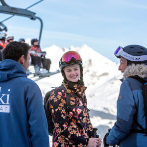 Ski Beat launches 2025-26 ski season! Three helmeted skiers chat on a snowy slope, with one wearing a Ski BEAT jacket, while others ride a chairlift against mountain peaks.