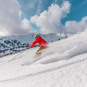 A skier in a bright red jacket carves through deep powder, sending up a spray of snow against a backdrop of snow-capped mountains and a partly cloudy blue sky.