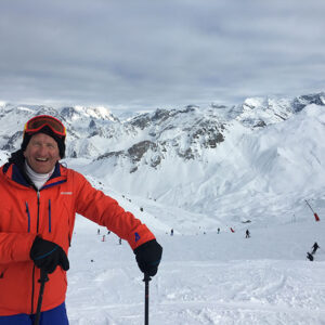 A grinning skier in an orange jacket and red goggles stands on a snowy slope with other skiers and rugged mountain peaks behind.